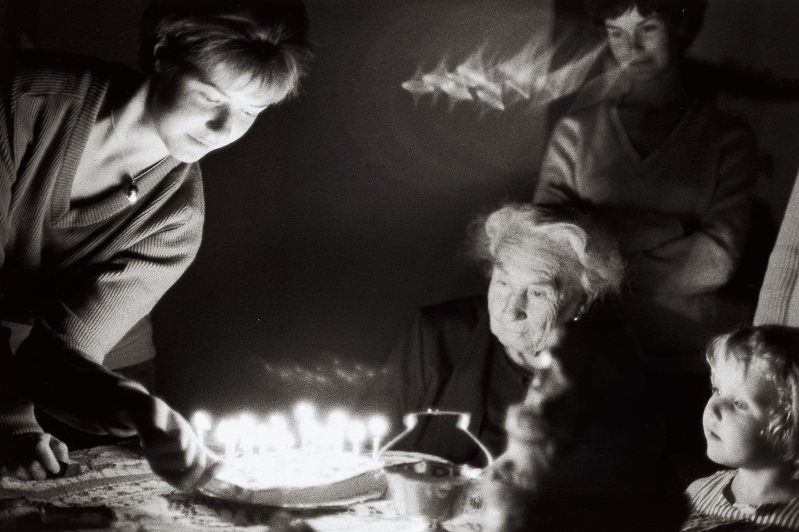 Black and white photograph, a family gathers to watch their grandmother blow out a candle.