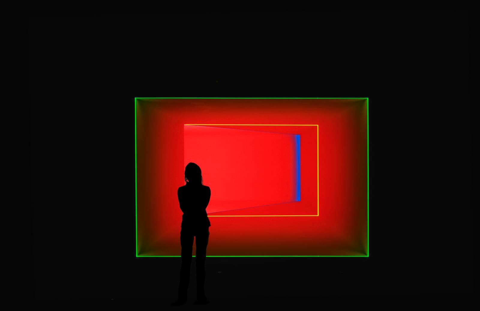 A figure is silhouetted against a bright red box outlined in green that appears to continue endlessly into the wall