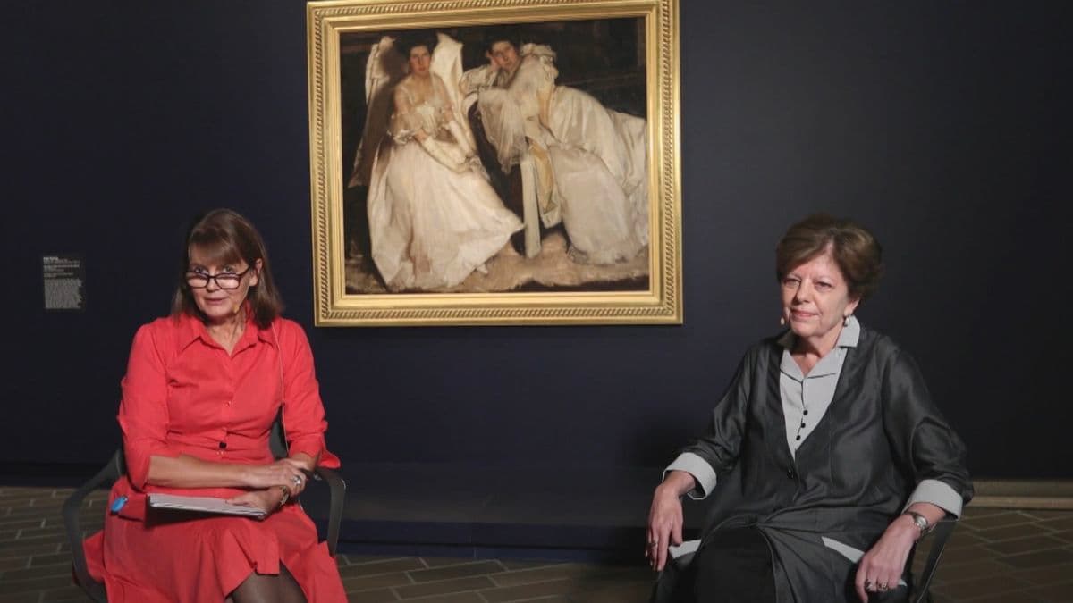 Video still of two women in conversation during an interview, seated in front painting in exhibition.