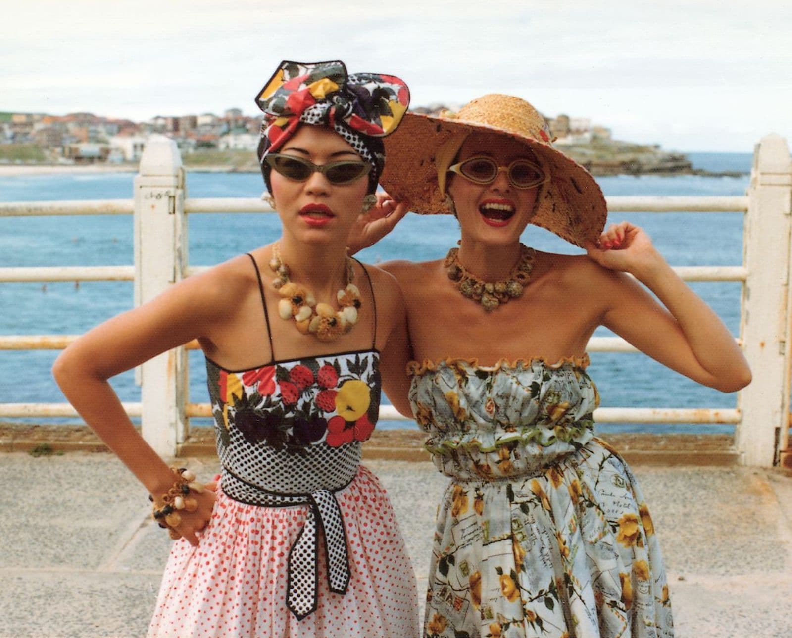 Two woman in bright hats, jewellery, sunglasses and dresses have their arms around each other, posing for a photograph